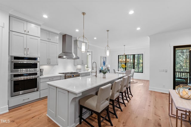 kitchen with a wealth of natural light, a center island with sink, wall chimney range hood, double oven, and light countertops