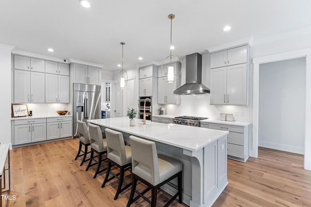 kitchen with a breakfast bar, appliances with stainless steel finishes, wall chimney exhaust hood, light wood finished floors, and light countertops