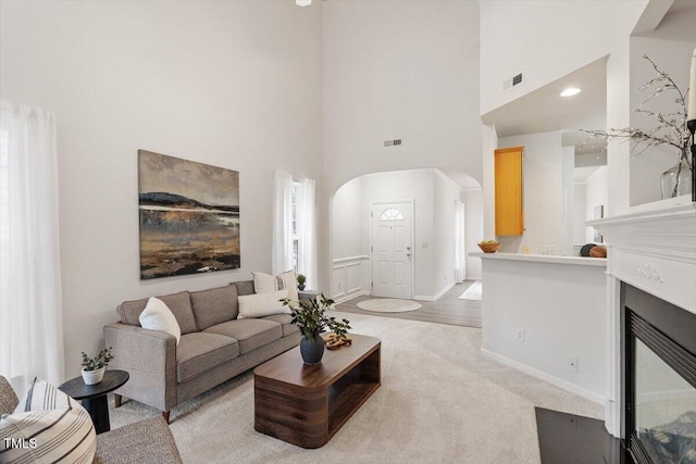 living room featuring visible vents, a fireplace with flush hearth, light carpet, a high ceiling, and arched walkways