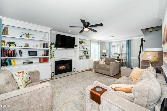 living room featuring built in features, wood finished floors, ceiling fan, a glass covered fireplace, and crown molding