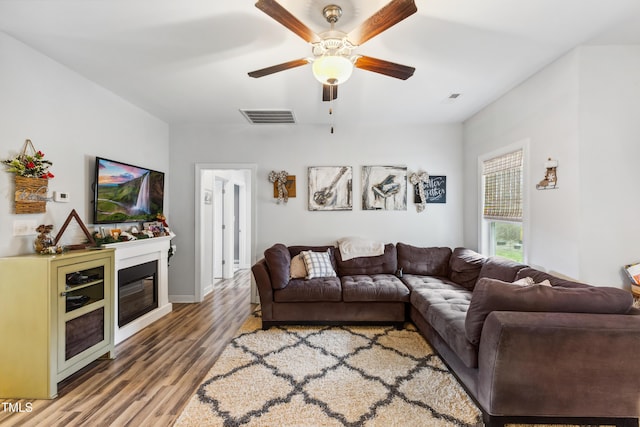 living room with visible vents, a ceiling fan, a glass covered fireplace, and wood finished floors