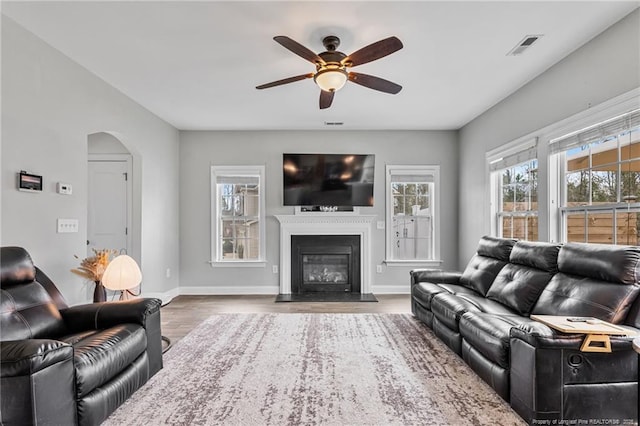 living area featuring a ceiling fan, wood finished floors, baseboards, arched walkways, and a glass covered fireplace