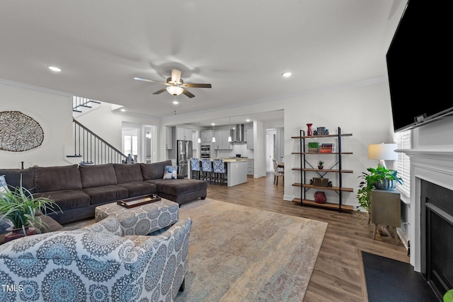 living area with crown molding, a fireplace with flush hearth, stairs, light wood-style floors, and a ceiling fan