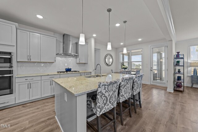 kitchen with light wood finished floors, stainless steel appliances, a sink, wall chimney range hood, and tasteful backsplash