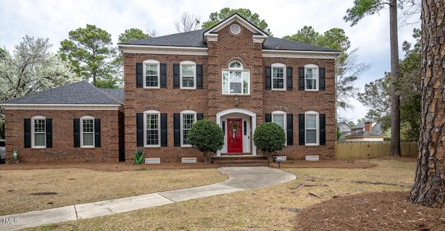 colonial-style house featuring fence, brick siding, and roof with shingles