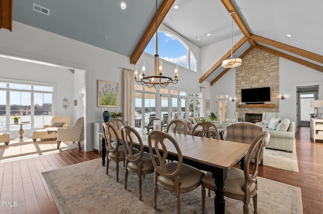 dining space featuring visible vents, dark wood-type flooring, a chandelier, beam ceiling, and a stone fireplace