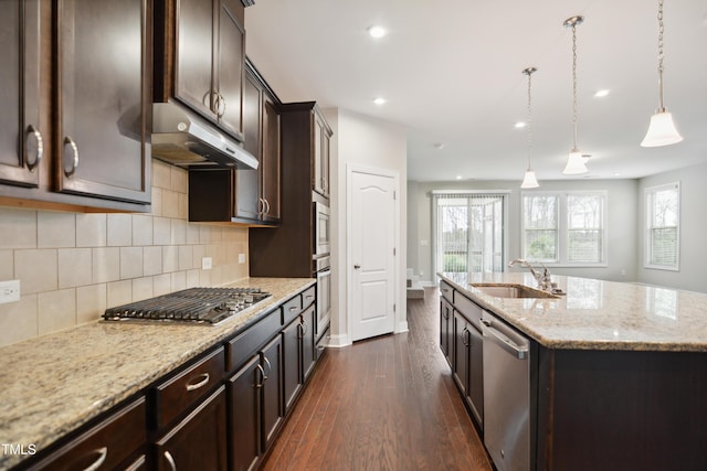 kitchen with dark brown cabinets, under cabinet range hood, a center island with sink, appliances with stainless steel finishes, and a sink