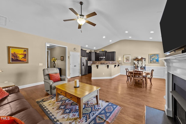 living area featuring a fireplace with flush hearth, vaulted ceiling, wainscoting, wood finished floors, and a ceiling fan