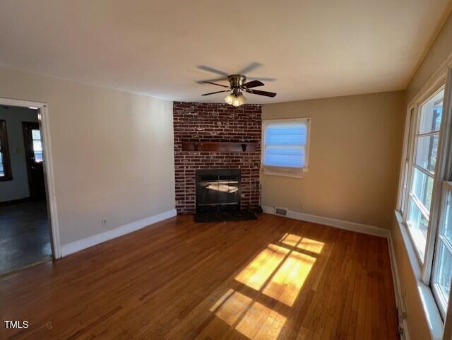 unfurnished living room with a ceiling fan, wood finished floors, visible vents, baseboards, and a brick fireplace