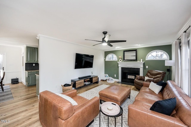 living area with baseboards, a fireplace with flush hearth, ceiling fan, ornamental molding, and light wood-type flooring