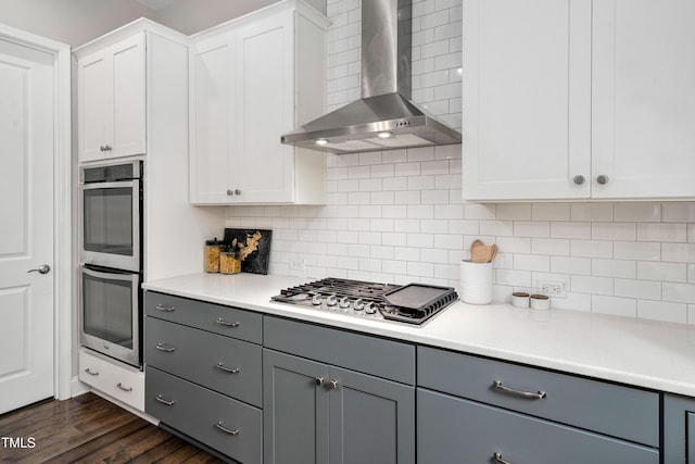 kitchen with dark wood-style floors, gray cabinets, stainless steel appliances, wall chimney exhaust hood, and backsplash