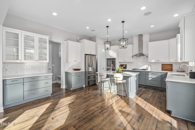 kitchen with a breakfast bar, an island with sink, appliances with stainless steel finishes, wall chimney exhaust hood, and a sink