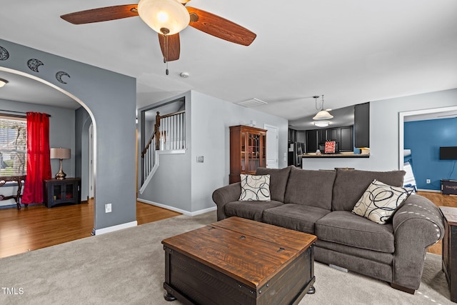 living area featuring light carpet, arched walkways, baseboards, ceiling fan, and stairs