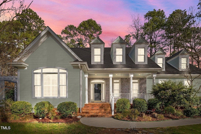 cape cod home with brick siding, a porch, and a shingled roof