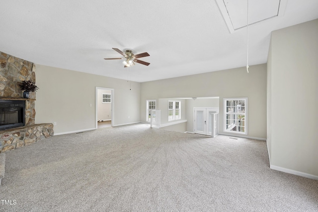 unfurnished living room featuring a stone fireplace, attic access, ceiling fan, and carpet flooring