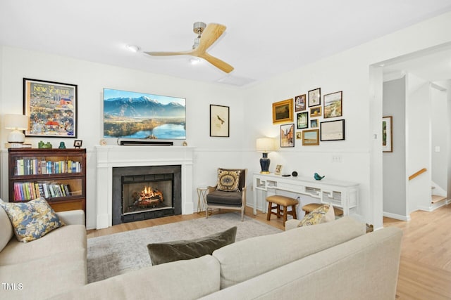 living area with a ceiling fan, baseboards, a warm lit fireplace, stairs, and light wood-type flooring