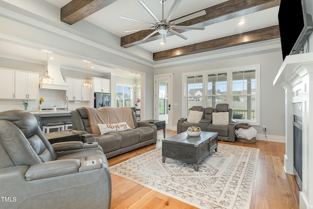 living area with beam ceiling, light wood-style flooring, ceiling fan with notable chandelier, a fireplace, and baseboards