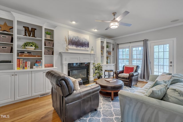 living room featuring a ceiling fan, visible vents, a fireplace with flush hearth, light wood-style floors, and crown molding