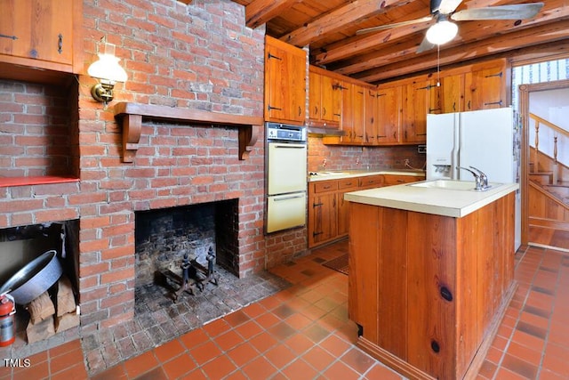 kitchen featuring white appliances, brown cabinetry, beam ceiling, light countertops, and a brick fireplace