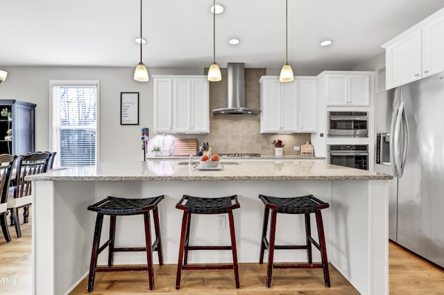 kitchen with backsplash, white cabinetry, stainless steel appliances, wall chimney exhaust hood, and light wood finished floors