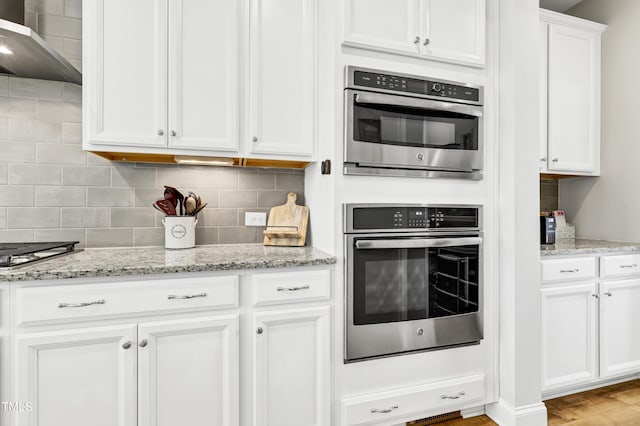 kitchen featuring light stone counters, backsplash, white cabinetry, double oven, and wall chimney exhaust hood