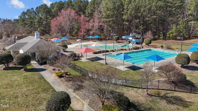 community pool with a patio, fence, a water slide, a yard, and a view of trees