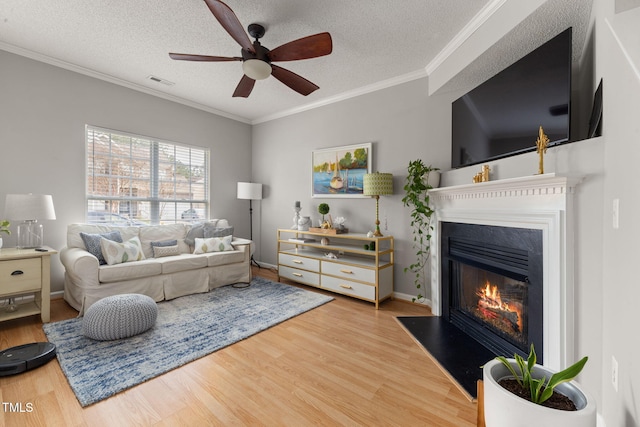 living area featuring a ceiling fan, wood finished floors, visible vents, a textured ceiling, and a glass covered fireplace