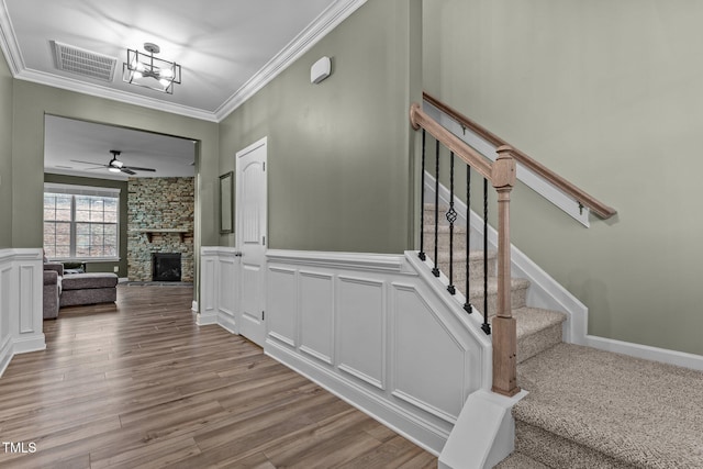 stairway featuring wood finished floors, visible vents, ornamental molding, a stone fireplace, and a decorative wall