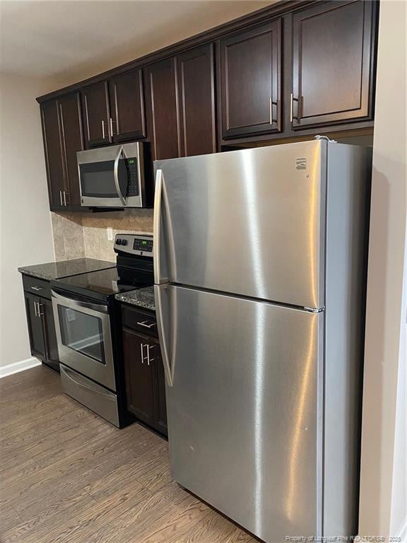 kitchen with dark stone countertops, stainless steel appliances, dark brown cabinetry, light wood-style floors, and backsplash