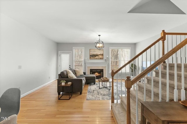 living area featuring baseboards, light wood-style flooring, stairs, a glass covered fireplace, and a notable chandelier