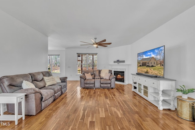 living area featuring baseboards, a lit fireplace, ceiling fan, and light wood finished floors