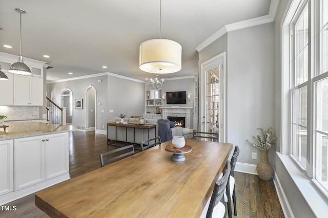 dining space featuring baseboards, dark wood-style flooring, a warm lit fireplace, arched walkways, and crown molding