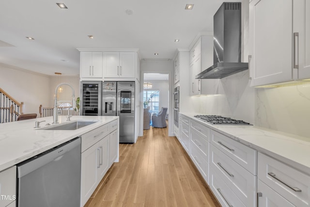 kitchen featuring crown molding, wall chimney range hood, appliances with stainless steel finishes, white cabinetry, and a sink