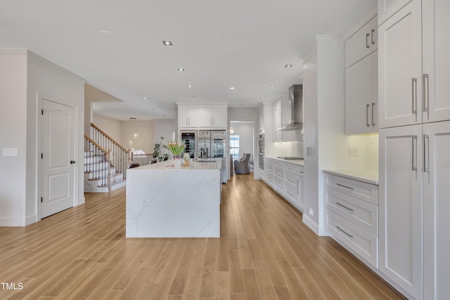 kitchen featuring wall chimney exhaust hood, an island with sink, light wood finished floors, and stainless steel appliances