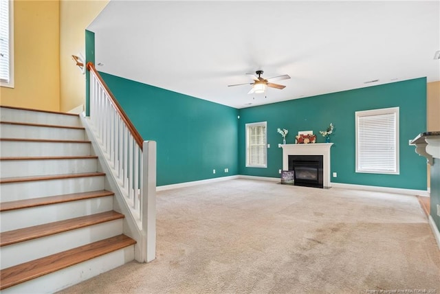 unfurnished living room with stairway, baseboards, a fireplace with flush hearth, ceiling fan, and carpet flooring