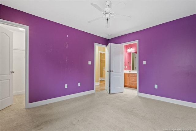 unfurnished bedroom featuring a walk in closet, baseboards, light colored carpet, ensuite bath, and a ceiling fan