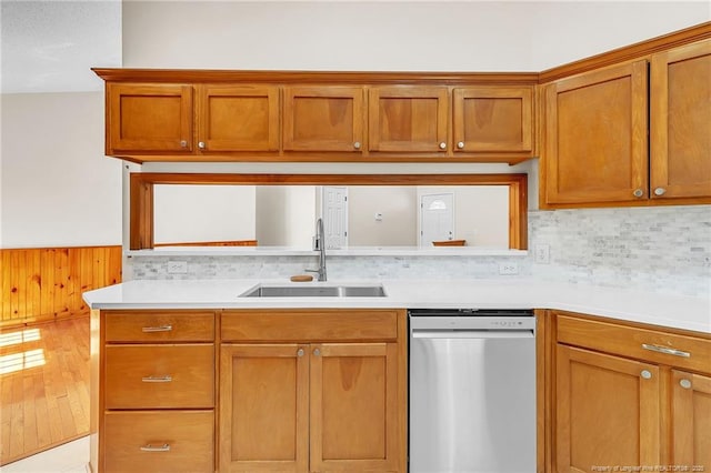 kitchen featuring light countertops, wainscoting, stainless steel dishwasher, brown cabinetry, and a sink