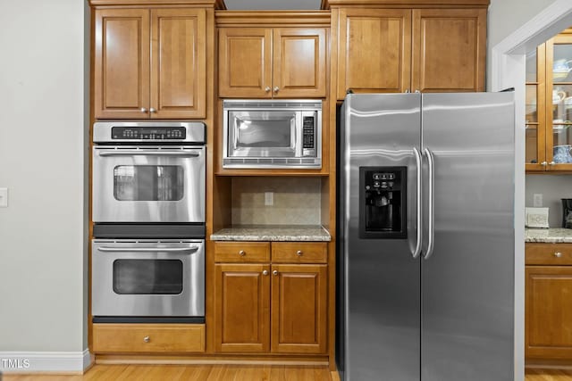 kitchen featuring tasteful backsplash, light wood-type flooring, light stone counters, brown cabinetry, and stainless steel appliances