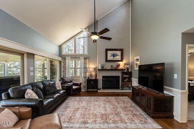 living room featuring high vaulted ceiling, wood finished floors, wainscoting, a fireplace, and ceiling fan