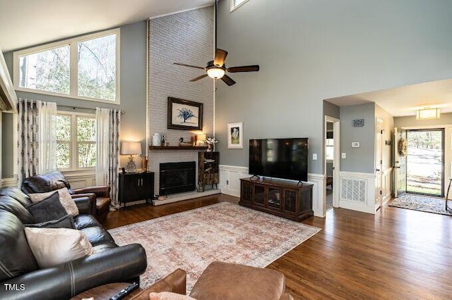 living area with ceiling fan, dark wood-style floors, wainscoting, a brick fireplace, and a towering ceiling