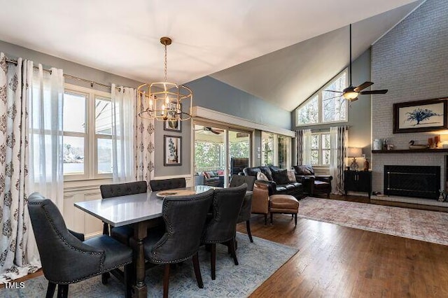 dining room with high vaulted ceiling, a brick fireplace, wood finished floors, and ceiling fan with notable chandelier