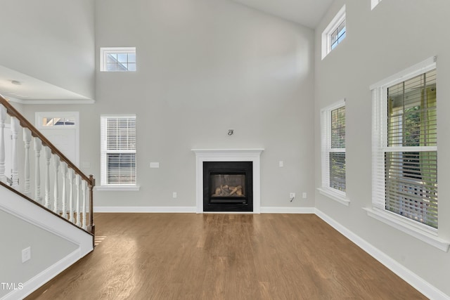 unfurnished living room featuring wood finished floors, baseboards, a high ceiling, stairs, and a glass covered fireplace