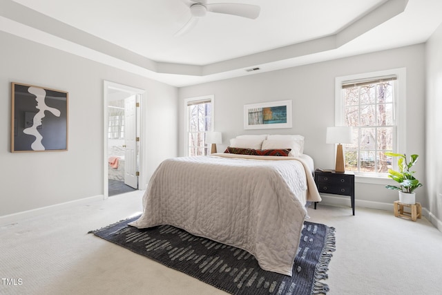 carpeted bedroom with a tray ceiling, multiple windows, baseboards, and ensuite bath
