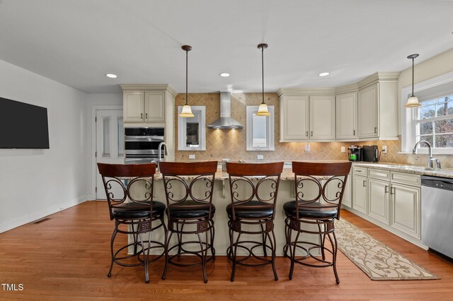 kitchen with light wood-type flooring, cream cabinetry, a sink, appliances with stainless steel finishes, and wall chimney range hood