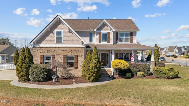 craftsman inspired home with fence, roof with shingles, covered porch, a front lawn, and brick siding