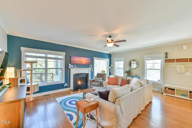 living room with light wood-type flooring, a fireplace with flush hearth, visible vents, ornamental molding, and ceiling fan