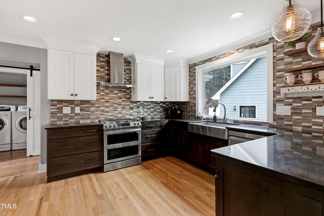 kitchen featuring washing machine and clothes dryer, dark countertops, wall chimney range hood, appliances with stainless steel finishes, and a sink