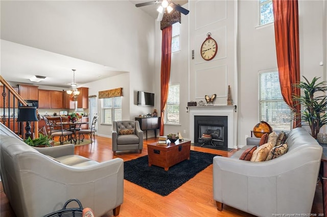 living area featuring stairs, ceiling fan with notable chandelier, a high ceiling, light wood-style flooring, and a fireplace