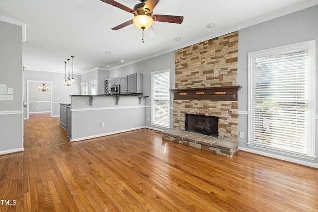 unfurnished living room featuring ceiling fan with notable chandelier, hardwood / wood-style floors, a stone fireplace, crown molding, and baseboards