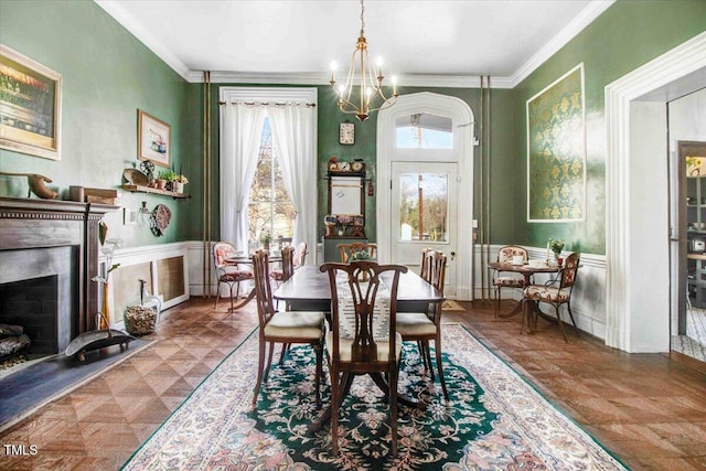 dining space featuring a fireplace with raised hearth, a decorative wall, an inviting chandelier, wainscoting, and crown molding